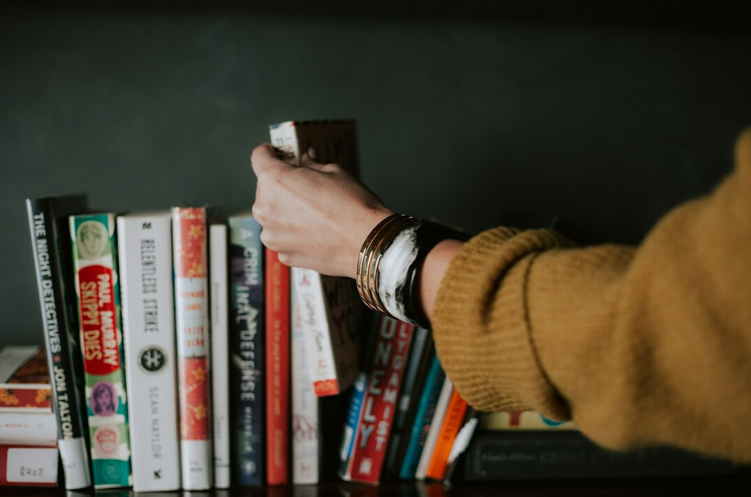 Person's hands reading an open book at a wooden desk with a lamp